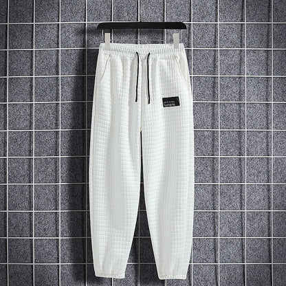 Pantaloni sportivi bianchi a quadri, con elastico in vita e coulisse nera, appesi su griglia metallica. Stile casual, moda uomo.