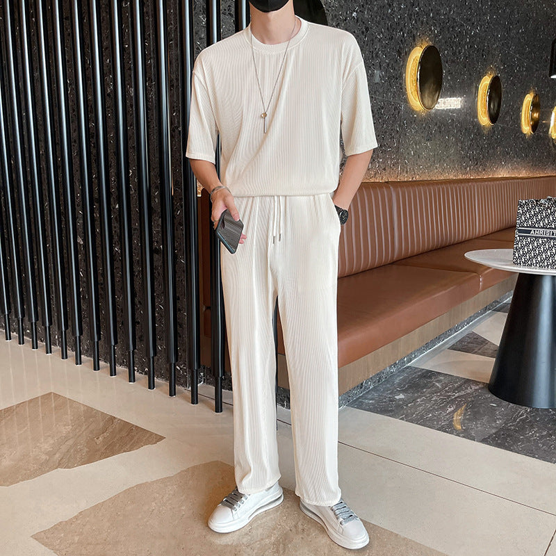 Uomo in abbigliamento casual bianco, con t-shirt e pantaloni coordinati, in un ambiente moderno con pareti scure e sedute in pelle marrone.
