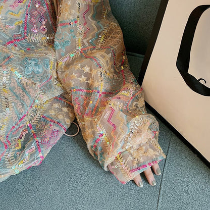 Abito in tulle ricamato con paillettes colorate su sfondo beige, dettagli geometrici e floreali, accanto a una borsa bianca su divano blu.