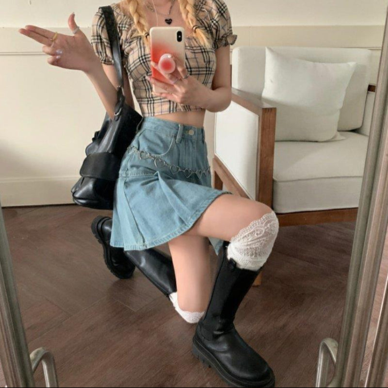 Giovane donna in posa con smartphone, indossa top a quadri, gonna di jeans, stivali neri e calze bianche. Stile moda casual femminile.