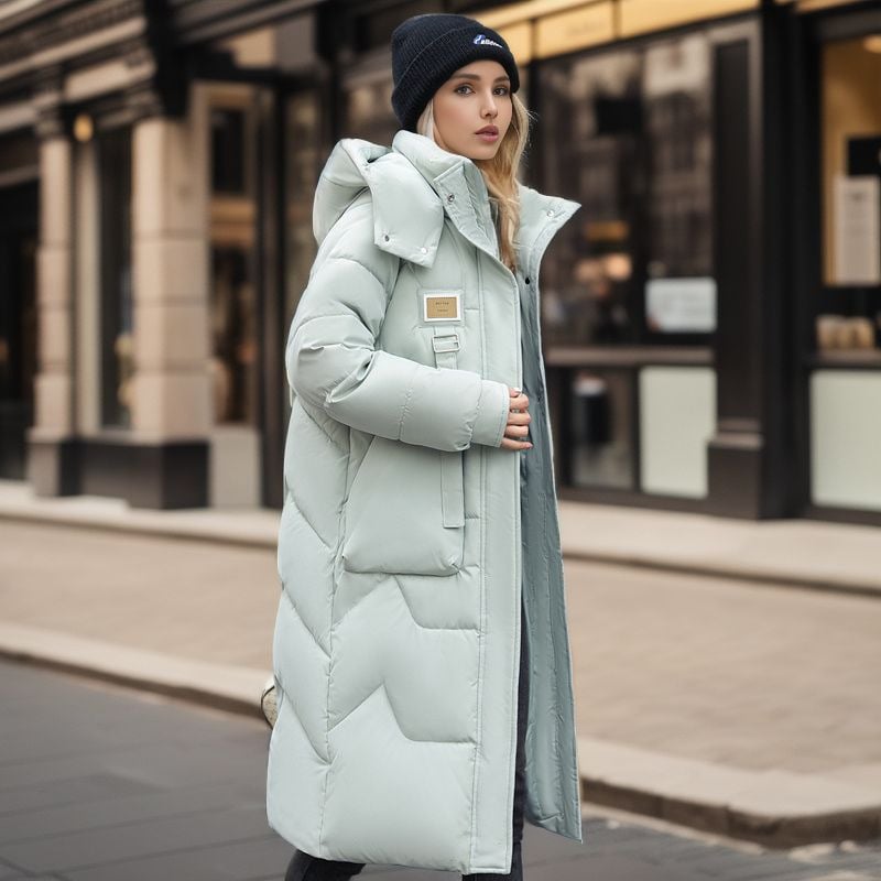 Donna con cappotto lungo imbottito verde chiaro e berretto nero cammina in città. Moda invernale, abbigliamento caldo, stile urbano elegante.