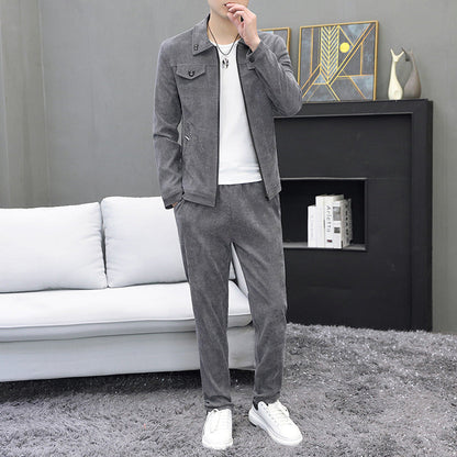 Uomo in abbigliamento casual grigio, giacca e pantaloni, su tappeto grigio. Sfondo con divano bianco e mensola con libri e decorazioni moderne.
