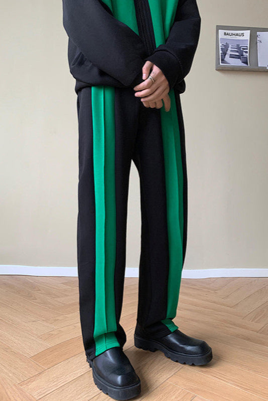 Pantaloni neri con strisce verdi, indossati con giacca nera e scarpe nere su pavimento in legno. Sfondo con parete beige e rivista Bauhaus.