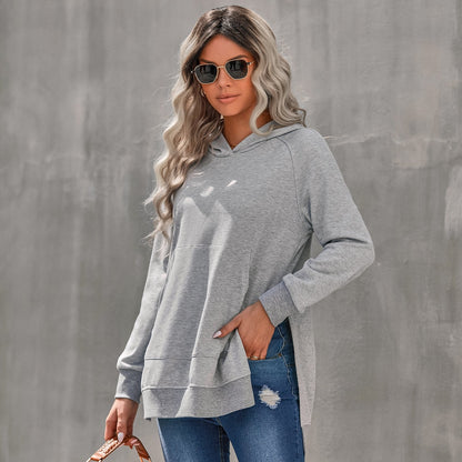 Donna con occhiali da sole indossa felpa grigia con cappuccio e jeans strappati, sfondo grigio. Moda casual, street style, abbigliamento autunnale.