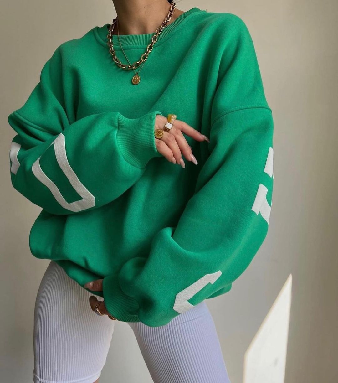 Donna con felpa verde oversize, dettagli bianchi sulle maniche, indossa collana e anelli dorati, pantaloni bianchi a coste. Moda casual chic.