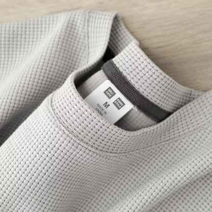 Maglia grigia a trama waffle, taglia media, con etichetta visibile su sfondo in legno chiaro. Tessuto casual e moderno, ideale per moda autunnale.