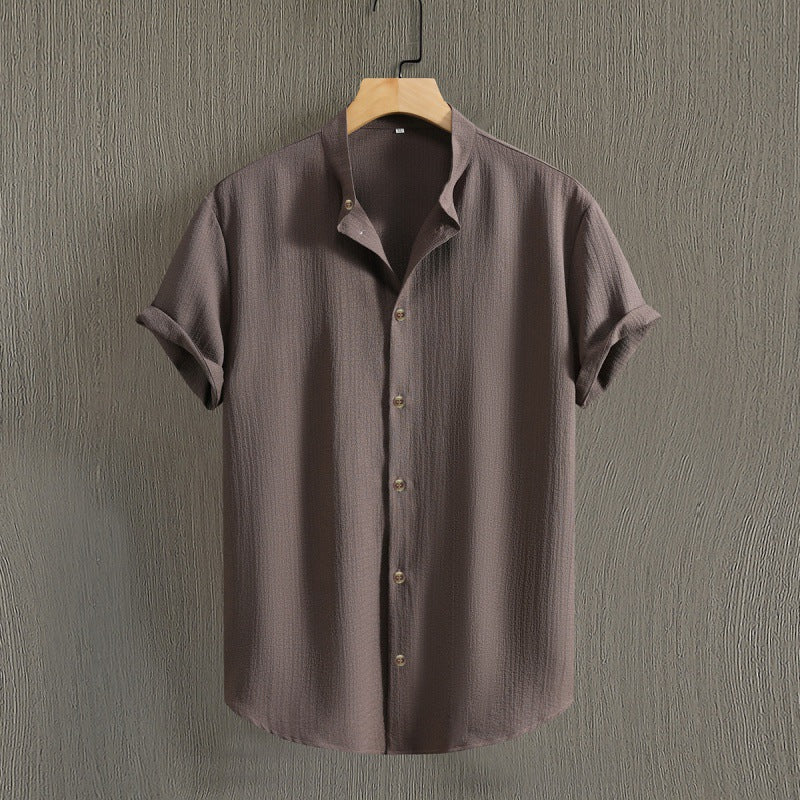 Camicia marrone a maniche corte, tessuto leggero, appesa su gruccia di legno, sfondo grigio. Moda uomo, abbigliamento estivo casual.