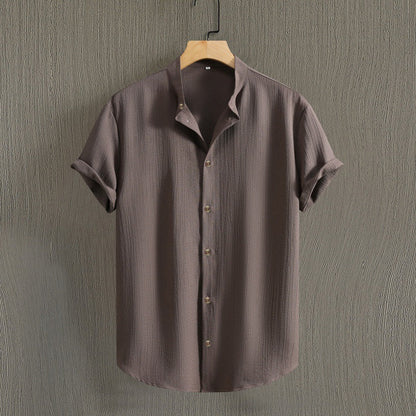 Camicia marrone a maniche corte, tessuto leggero, appesa su gruccia di legno, sfondo grigio. Moda uomo, abbigliamento estivo casual.