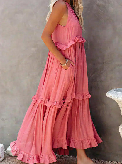 Donna in abito lungo rosa con balze, stile boho chic, posa di profilo su sfondo grigio. Moda estiva, abbigliamento femminile elegante.