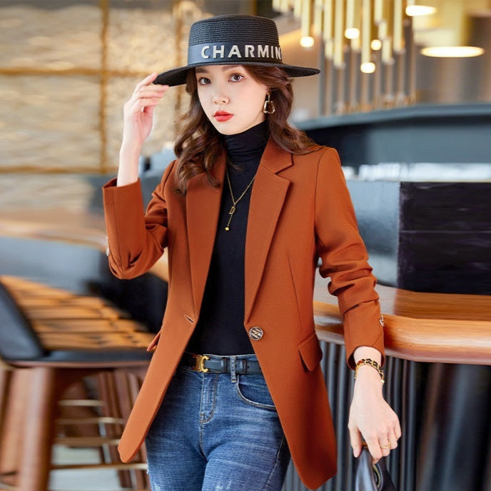 Donna con cappello nero "Charmin", blazer marrone, maglia nera e jeans, in un ambiente moderno. Moda autunnale elegante e casual.