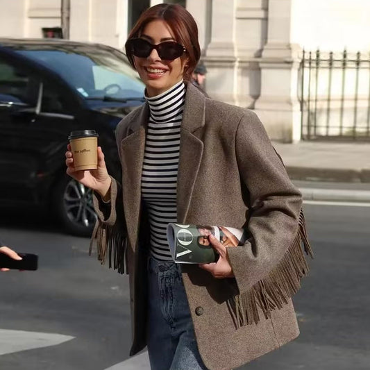 Donna sorridente con occhiali da sole, indossa cappotto con frange e maglia a righe, tiene caffè e rivista. Moda autunnale urbana, street style.