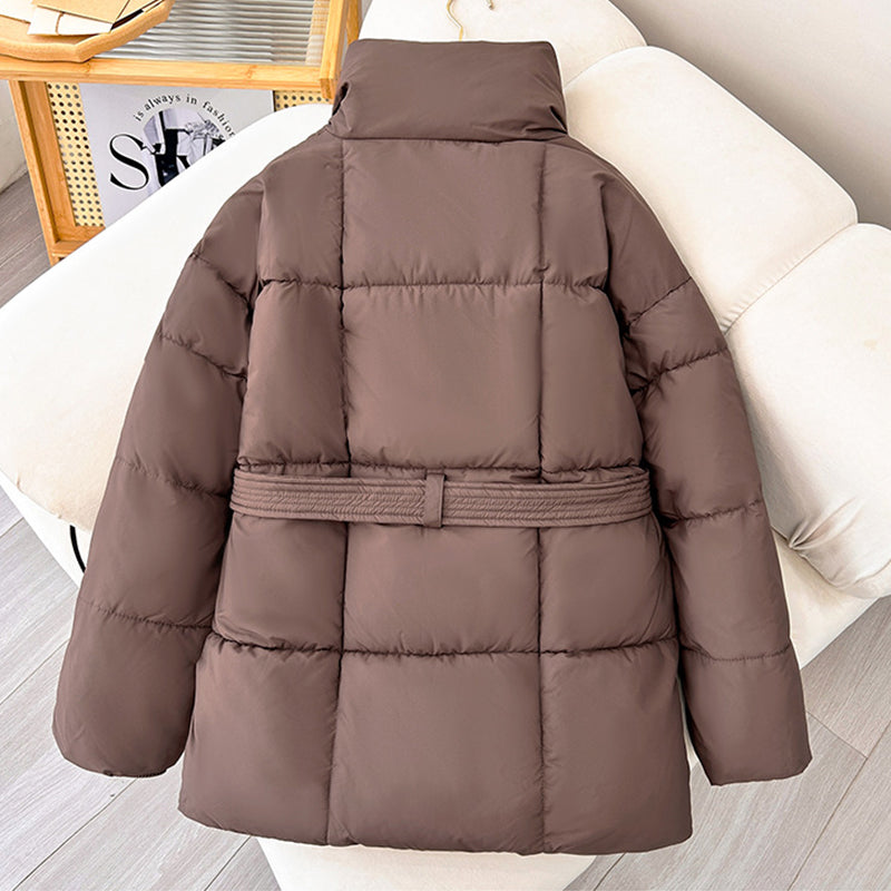 Giacca invernale imbottita marrone su divano beige, design trapuntato, cintura in vita, ideale per moda autunno-inverno, abbigliamento caldo donna.