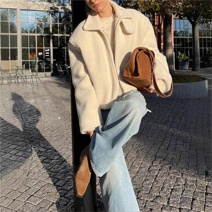 Giacca in lana bianca oversize, jeans a zampa, borsa marrone, stivali beige, sfondo urbano con vetrate e sedie all'aperto. Moda autunnale casual.