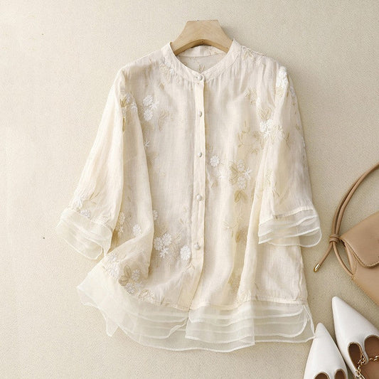 Camicia donna in lino bianco con ricami floreali, maniche a tre quarti, abbinata a borsa beige e scarpe bianche eleganti su sfondo chiaro.