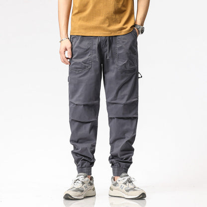 Pantaloni cargo grigi da uomo, stile casual, con tasche multiple, indossati con scarpe sportive New Balance e maglietta marrone.