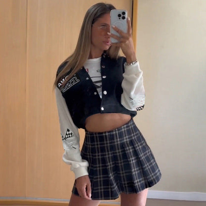 Giovane donna in posa davanti a uno specchio, indossa una giacca sportiva nera e bianca e una gonna a quadri. Scatta un selfie con uno smartphone.
