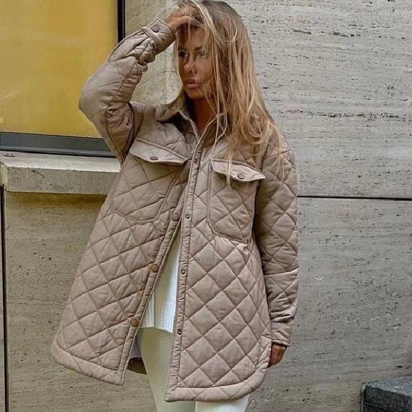 Donna con cappotto trapuntato beige, posa davanti a un muro di cemento. Moda autunno, stile urbano, abbigliamento casual chic.