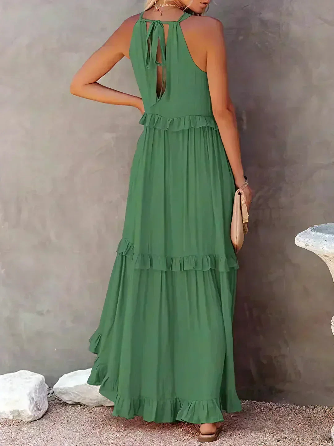 Donna in abito lungo verde con balze, schiena scoperta e sandali, posa di lato su sfondo grigio. Moda estiva elegante e casual.