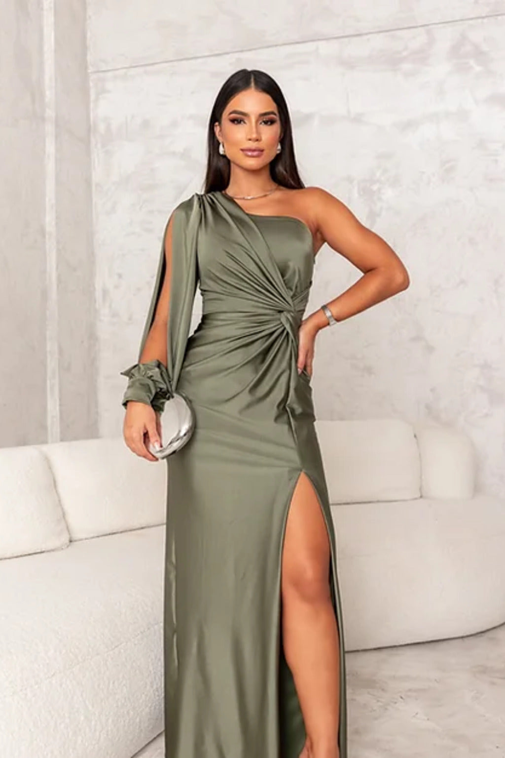 Donna in abito lungo verde oliva con spacco, seduta su divano bianco. Moda elegante, abbigliamento da sera, stile sofisticato.
