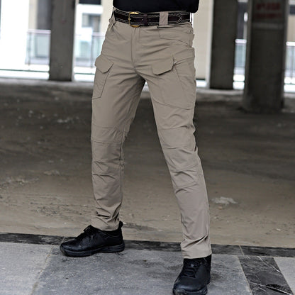 Pantaloni tattici beige con tasche multiple, indossati con cintura nera e scarpe nere, in un ambiente industriale.