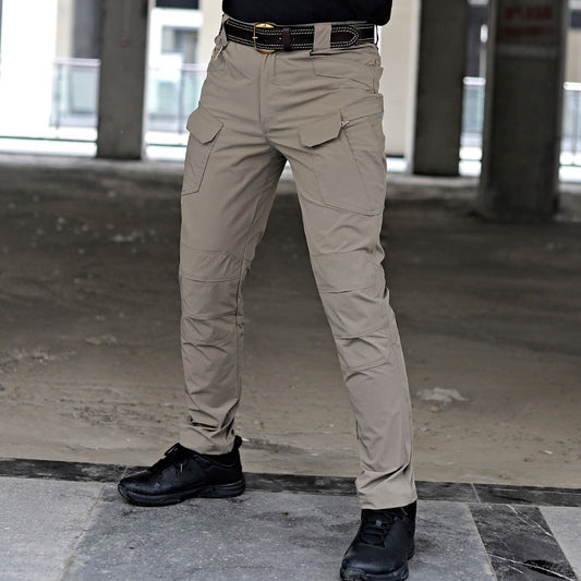 Pantaloni tattici beige con tasche multiple, indossati con cintura nera e scarpe nere, in un ambiente industriale.