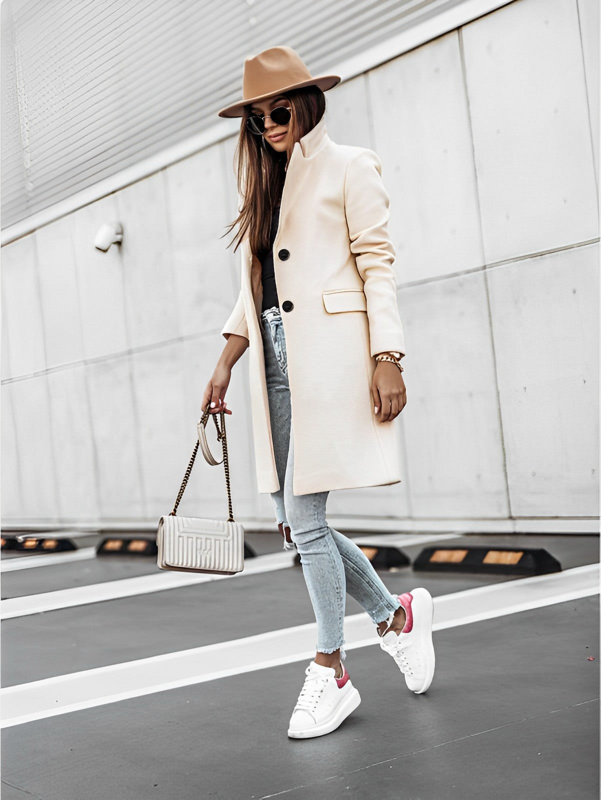 Donna alla moda con cappotto beige, jeans strappati, sneakers bianche e cappello marrone, cammina su strada urbana. Stile autunnale elegante.
