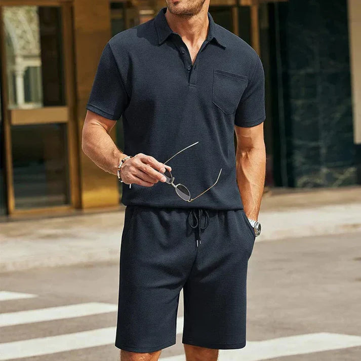 Uomo in abbigliamento casual estivo, indossa polo e pantaloncini blu scuro, tiene occhiali da sole. Moda maschile, look estivo elegante.
