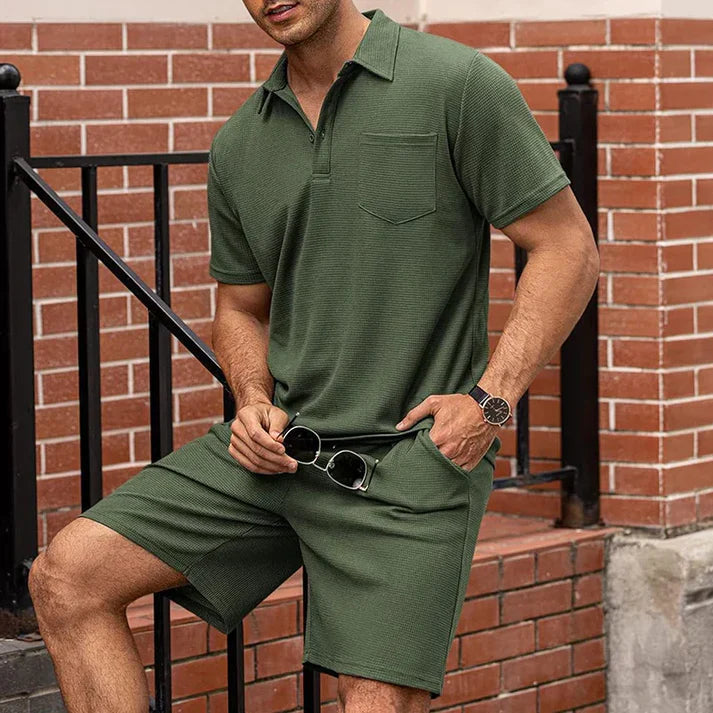 Uomo in completo estivo verde oliva, polo e pantaloncini, con orologio nero e occhiali da sole. Moda casual maschile, abbigliamento estivo elegante.