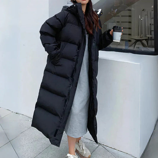 Donna con cappotto lungo nero imbottito, abito grigio e scarpe da ginnastica bianche, tiene una tazza da caffè. Moda invernale casual e moderna.