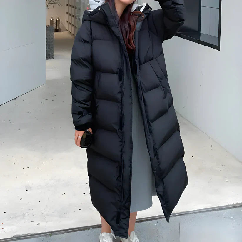 Donna con cappotto lungo nero imbottito, moda invernale, abbigliamento caldo, stile casual, scarpe sportive bianche, ambiente urbano minimalista.