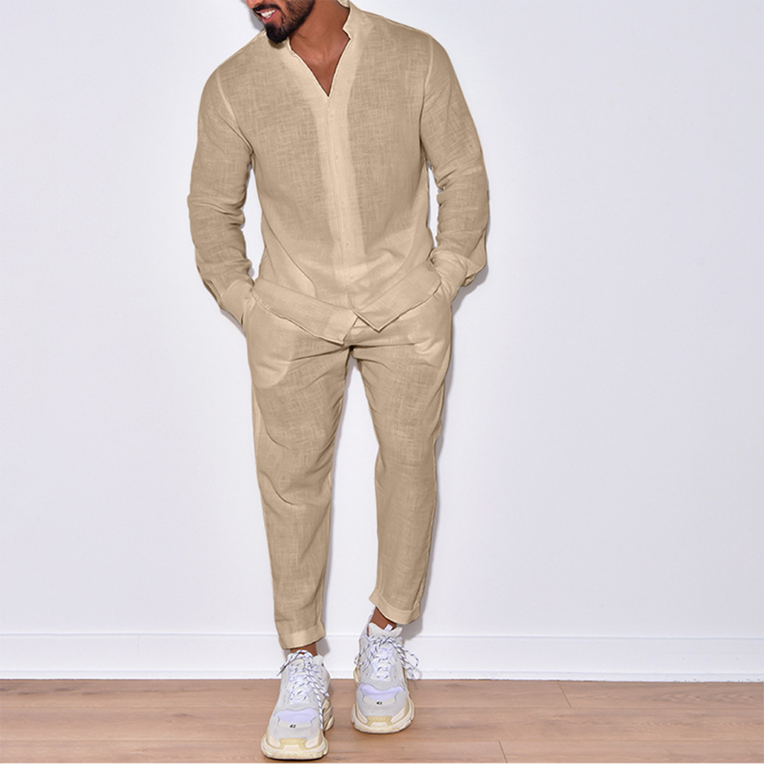 Uomo in abito di lino beige, camicia e pantaloni coordinati, scarpe da ginnastica bianche, posa su pavimento in legno, moda casual elegante.