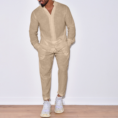 Uomo in abito di lino beige, camicia e pantaloni coordinati, scarpe da ginnastica bianche, posa su pavimento in legno, moda casual elegante.