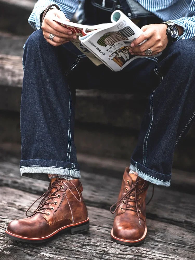 Uomo seduto su gradini indossa stivali in pelle marrone, jeans e camicia a righe, mentre legge una rivista. Moda casual maschile, calzature eleganti.