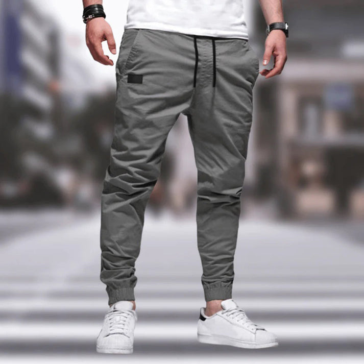 Uomo con pantaloni jogger grigi e scarpe da ginnastica bianche su sfondo urbano sfocato. Moda casual maschile, streetwear moderno.
