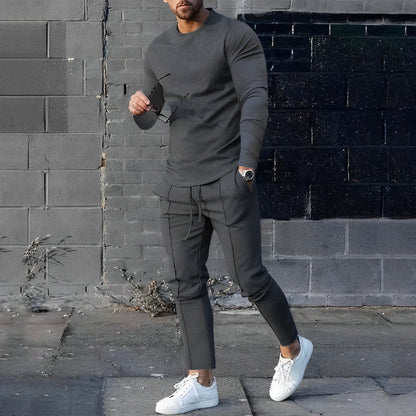 Uomo in abbigliamento casual grigio, con occhiali da sole e orologio, posa davanti a un muro di mattoni. Indossa scarpe da ginnastica bianche.