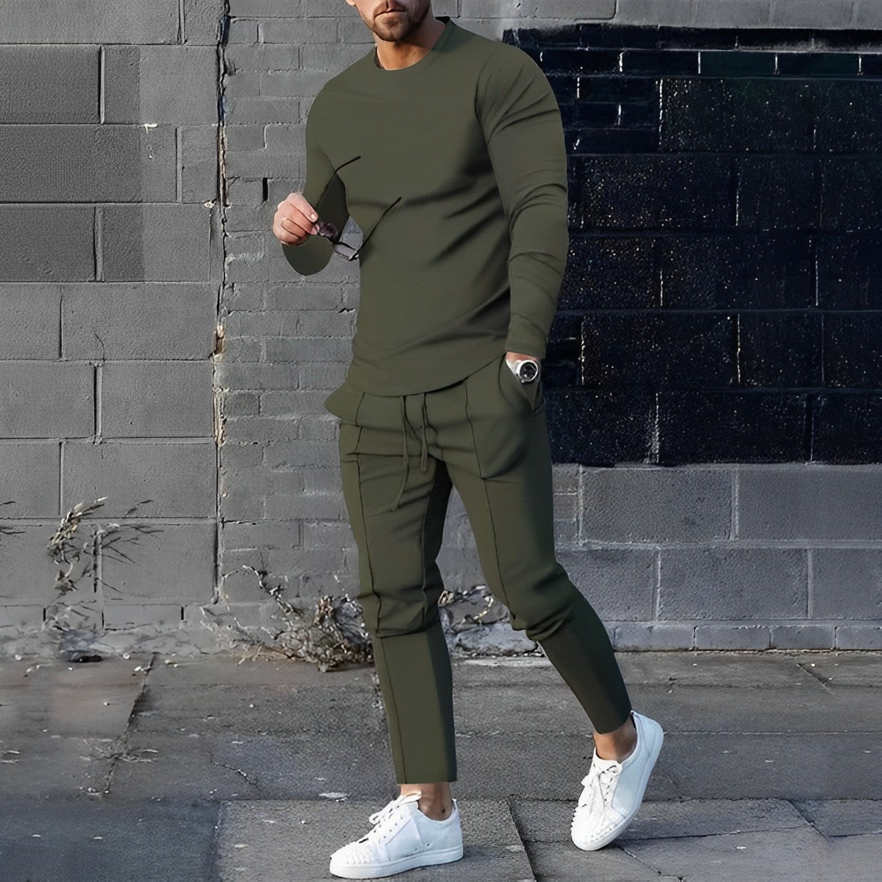 Uomo in abbigliamento casual verde oliva, con occhiali da sole e scarpe bianche, posa davanti a un muro di mattoni grigi e neri.