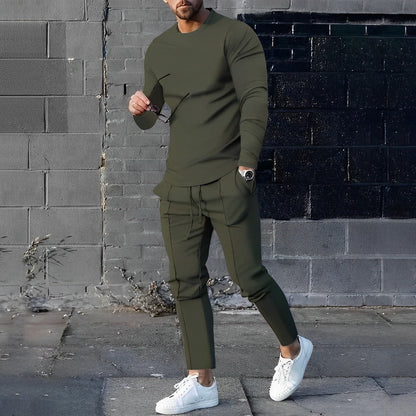 Uomo in abbigliamento casual verde oliva, con occhiali da sole e scarpe bianche, posa davanti a un muro di mattoni grigi e neri.