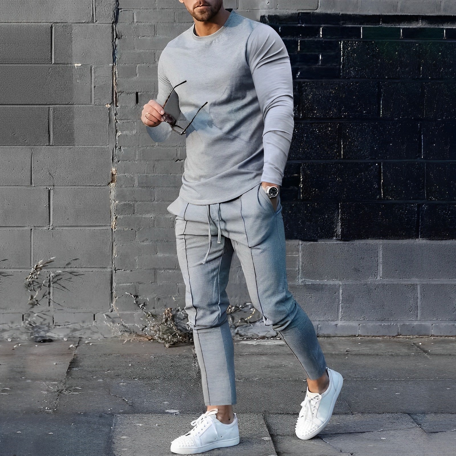 Uomo in abbigliamento casual grigio, con occhiali da sole in mano, posa davanti a un muro nero e grigio. Indossa scarpe bianche.