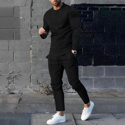 Uomo in abbigliamento casual nero con occhiali da sole e orologio, posa davanti a un muro di mattoni grigi, indossa scarpe bianche.