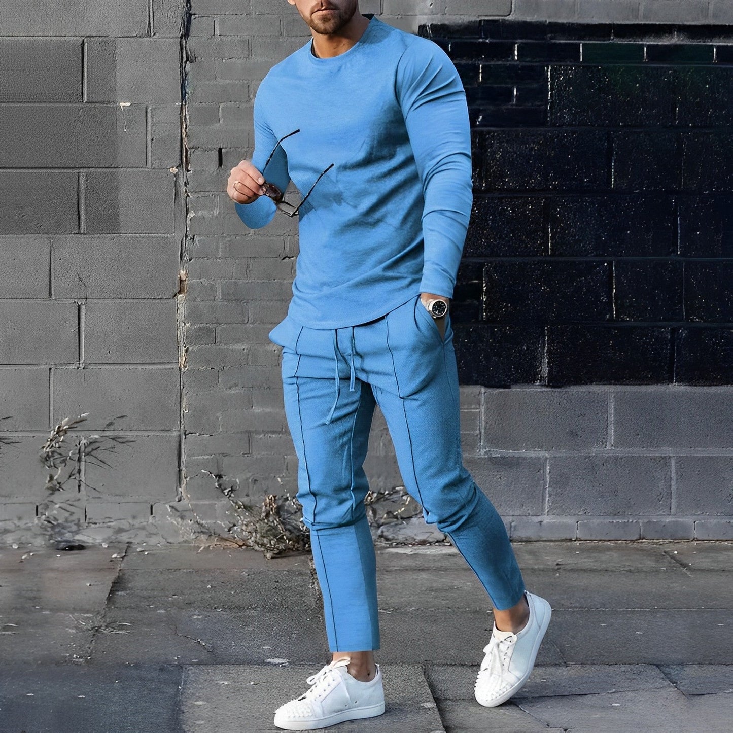 Uomo in tuta sportiva blu, cammina su marciapiede con occhiali da sole in mano. Indossa scarpe bianche. Sfondo di muro grigio e nero.