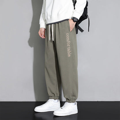 Pantaloni jogger grigi da uomo con scritta "comfortable", abbinati a felpa bianca e scarpe da ginnastica bianche, moda casual e sportiva.
