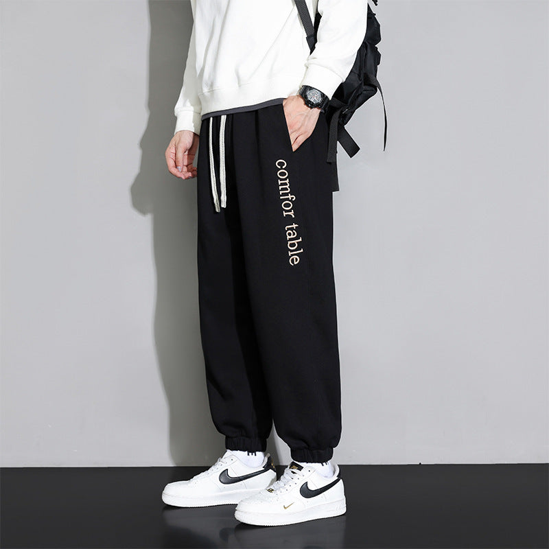 Pantaloni neri da uomo con scritta "comfortable", abbinati a felpa bianca e scarpe Nike bianche. Look casual e moderno, moda streetwear.