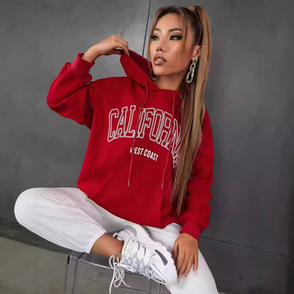 Donna con felpa rossa "California" e pantaloni bianchi, seduta su sfondo grigio. Moda casual, streetwear, stile urbano, abbigliamento trendy.