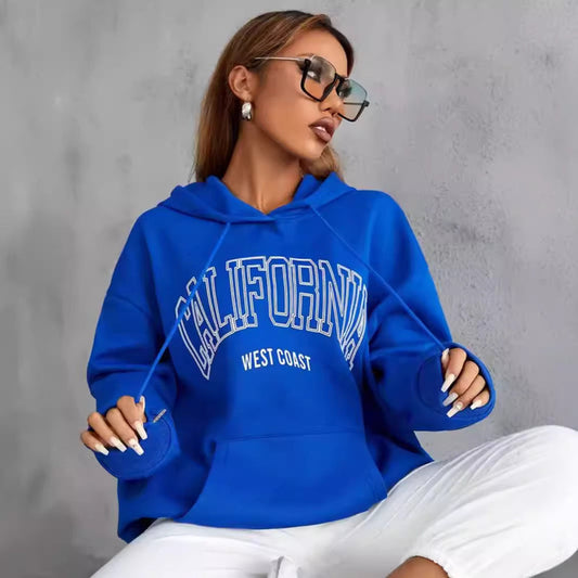 Donna con occhiali da sole indossa felpa blu con scritta "California West Coast", posa contro muro grigio. Moda casual, stile urbano, abbigliamento trendy.