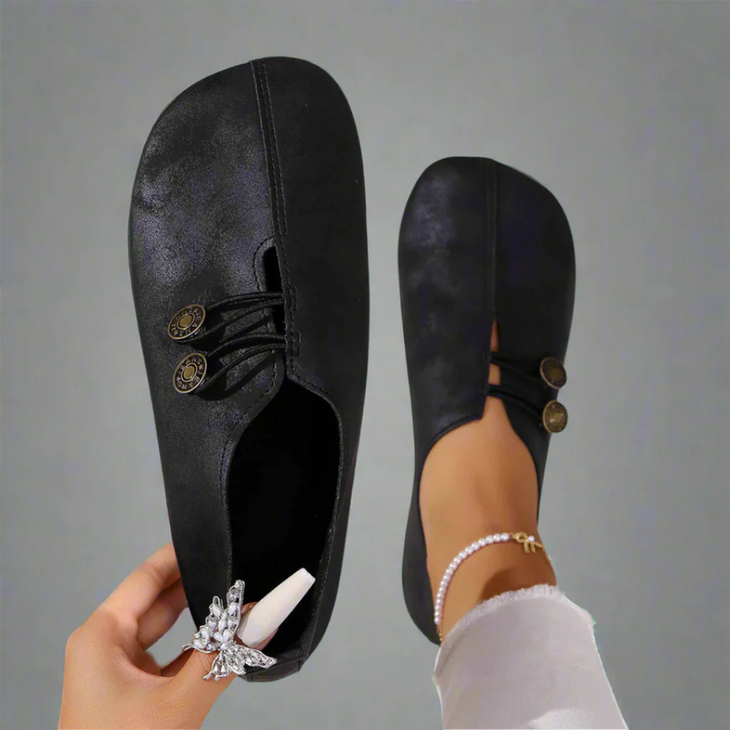 Scarpe da donna nere eleganti, design minimalista, chiusura con bottoni decorativi, indossate su sfondo grigio. Moda calzature femminili.