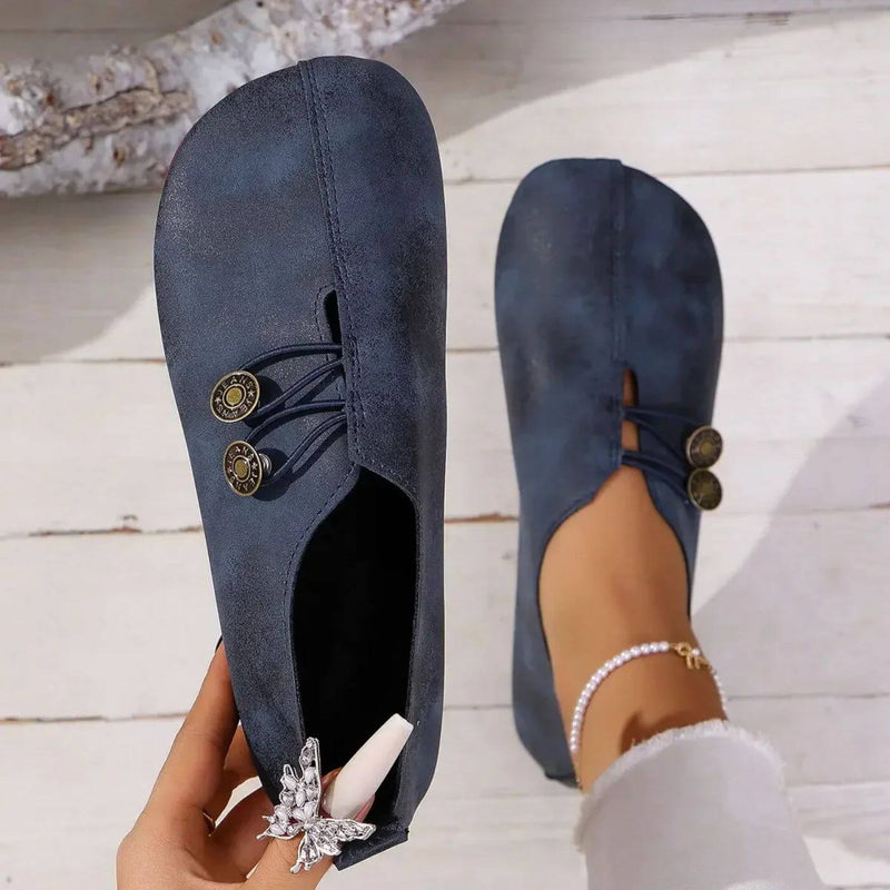 Scarpe da donna blu scuro in stile casual, con bottoni decorativi e design minimalista, indossate su pavimento in legno chiaro.