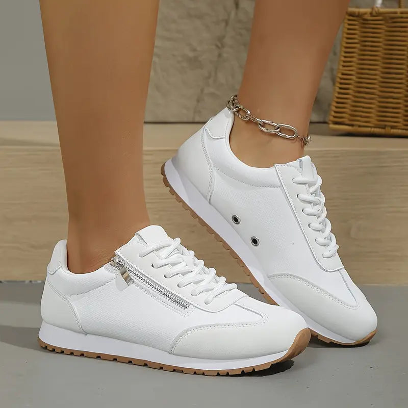 Scarpe da ginnastica bianche da donna con cerniera laterale, suola in gomma marrone, indossate su pavimento in legno. Accessorio cavigliera in metallo.