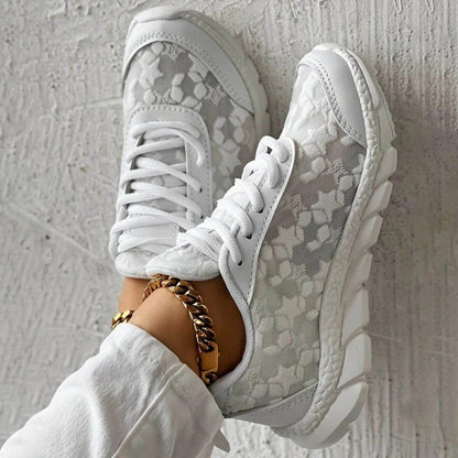 Scarpe da ginnastica bianche con motivo floreale, suola robusta e lacci, indossate con pantaloni bianchi e cavigliera dorata. Moda casual elegante.