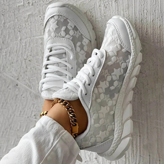 Scarpe da ginnastica bianche con motivo floreale, suola robusta e lacci, indossate con pantaloni bianchi e cavigliera dorata. Moda casual elegante.