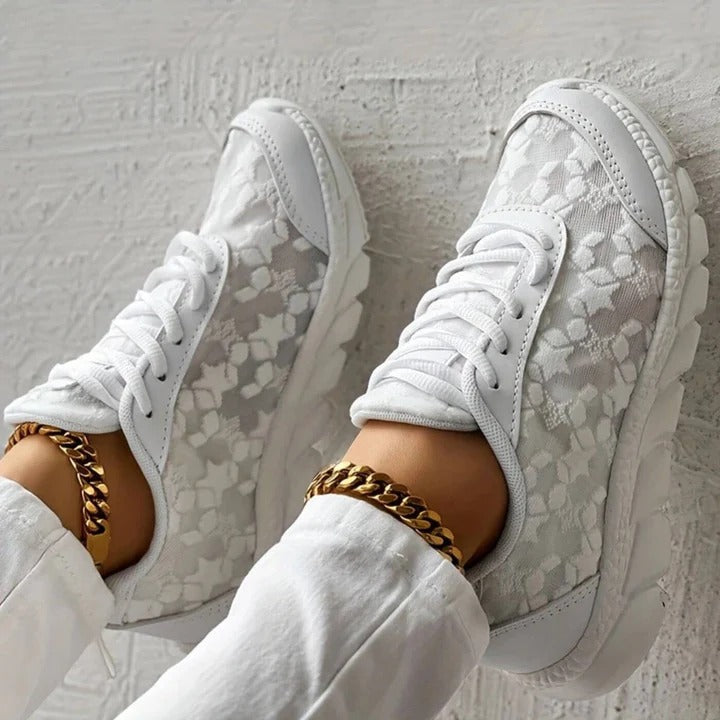 Scarpe da ginnastica bianche in pizzo floreale su sfondo bianco, indossate con pantaloni bianchi e cavigliere dorate. Moda casual elegante.